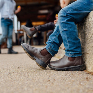 Close-up of Blundstone Boots 630 Series on a person sitting against a stone wall with a child and shed in the background.