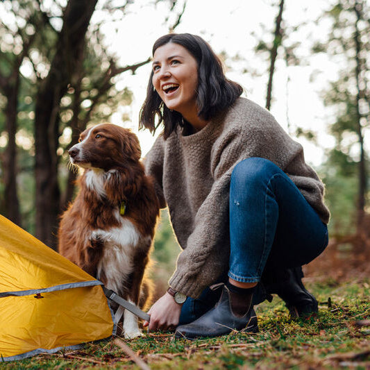 Woman and dog by a yellow tent in a forest wearing Blundstone Boots 600 Series