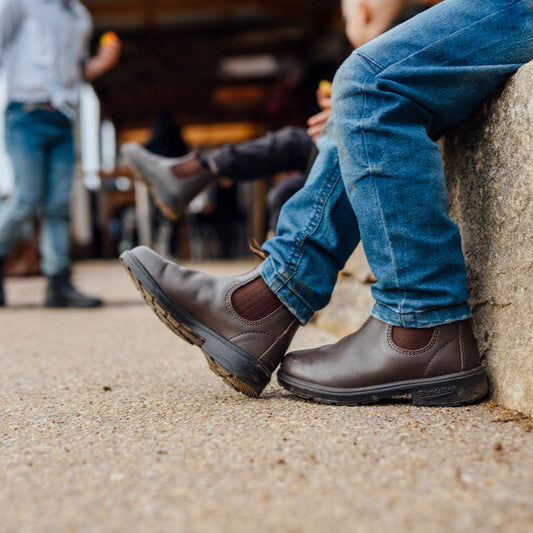 Close-up of Blundstone Boots 630 Series on a person sitting against a stone wall with a child and shed in the background.